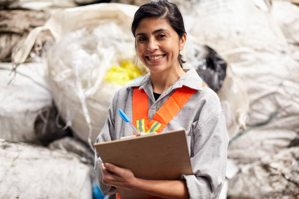 Cheerful latin female recycling coordinator looking at the camera while writing on clipboard stock photo