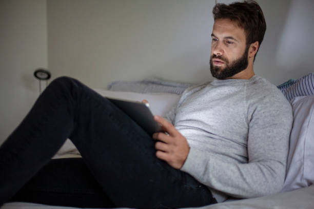 istock Young adult man lying on bed while watching something on digital tablet 1352094068
