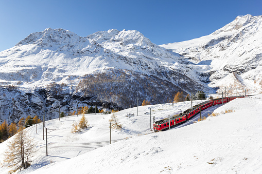 Alp Grum, Switzerland - November 05. 2021 : Red train from Rhaetian Railway is passing the train tracks with tight 180° curve at high Alp Grum. The Piz Palu peak is at the background.