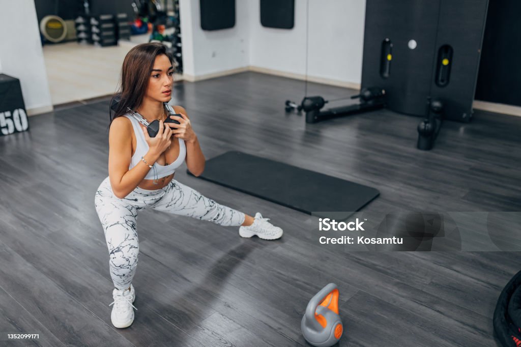 Holding weights in her hands, a young woman exercises in the gym. It transfers weight to one leg and thus activates all the muscles in the body A concentrated fit woman who trains in a modern gym. She is motivated to progress quickly 20-24 Years Stock Photo