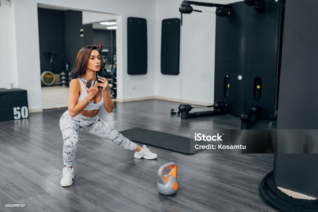 A young female athlete does leg exercises in the gym. She holds weights in hands and thus creates a greater effort with which she will progress faster A focused woman who exercises in the gym and prepares for a gym competition 20-24 Years Stock Photo