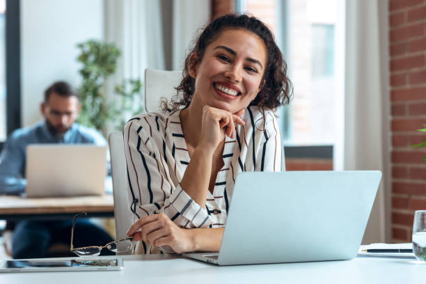 istock Smiling business woman working with laptop while looking at camera in modern startup office. 1352100252