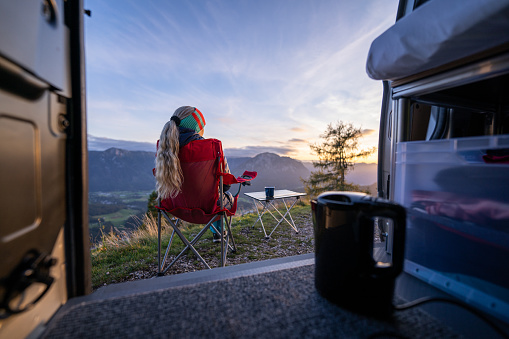 View From Inside Camper Van at Woman With Long Pigtail Sitting in Camping Chair Watching Sunset high up in the Mountains on Her Roadtrip Through Austria