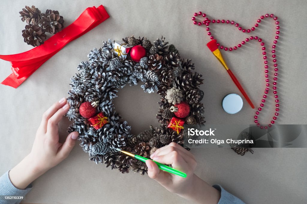 DIY. Step 3 top view of florist's hands with brush, Christmas wreath of fir cones, decorative toys Christmas Ornament Stock Photo