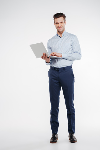 Vertical image of Smiling business man which standing in studio with laptop in hand and looking at camera. Isolated white background