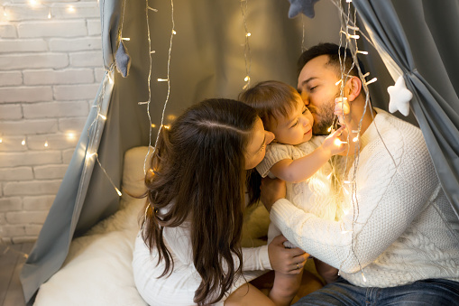 Parents kiss their daughter sitting under a New Year's garland of lights that interested their little daughter. The child holds the light bulbs of the glowing garland in his hands. Love and caring