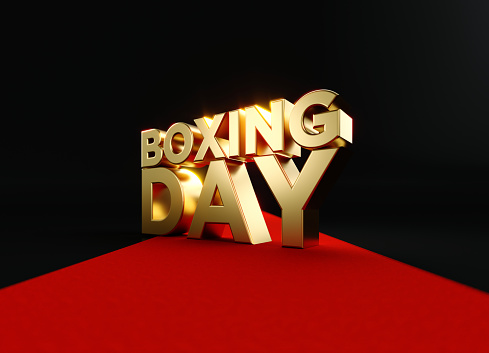 Gold colored Boxing Day sitting on red carpet before black background. Horizontal composition with copy space. Front view. Great use for Boxing Day concepts.