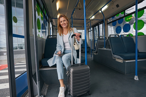 Smiling happy elegant female passenger with trolley suitcase sitting in transport vehicle