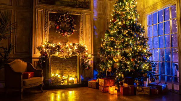 Photo of Christmas and New Year interior decoration. Green tree decorated with toys, gifts, present boxes, flashing garland, illuminated lamps. Fireplace and xmas tree. Cozy Christmas atmosphere