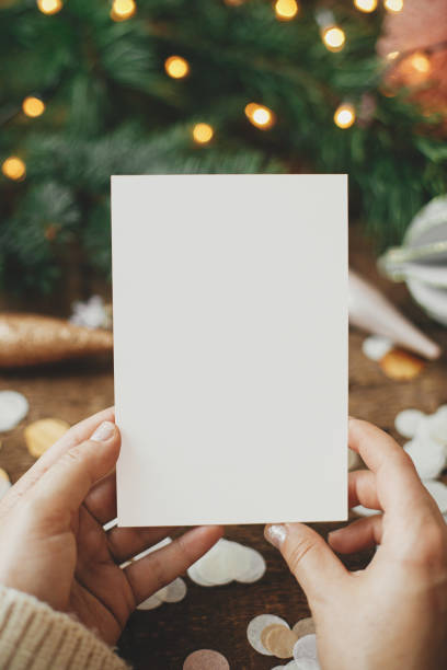 istock Hands holding empty greeting card and christmas ornaments, confetti, pine branches and lights on rustic wooden background. Christmas card mock up. Space for text. Seasons greetings. Template 1352124854