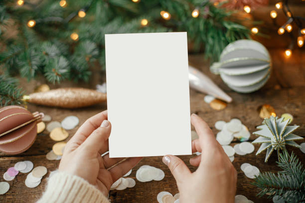 istock Hands holding empty greeting card and christmas ornaments, confetti, pine branches and lights on rustic wooden background. Christmas card mock up. Space for text. Seasons greetings. Template 1352126252