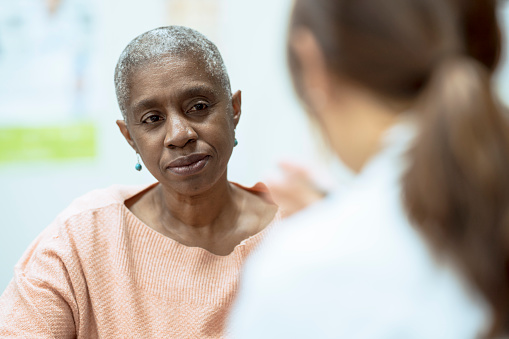 A senior woman with Cancer sits across from her doctor as they talk and discuss her treatment plan.  She is dressed casually and has a somber expression on her face.