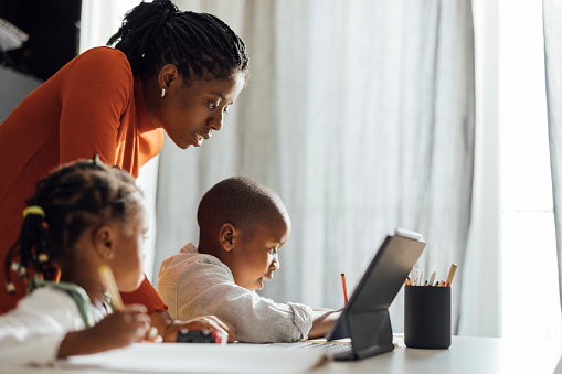 Brother and sister sitting at desk with their mother and writing their homework using a pen and a notebook in front of a digital tablet at home