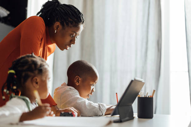 Serious Mother Helps her Children To Do Their Homework in a Living Room Brother and sister sitting at desk with their mother and writing their homework using a pen and a notebook in front of a digital tablet at home home schooling homework computer learning stock pictures, royalty-free photos & images