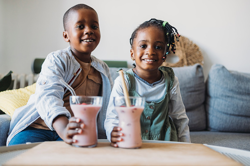 Cheerful smiling brother and sister holding a strawberry smoothie in hands while sitting on a sofa and looking at camera
