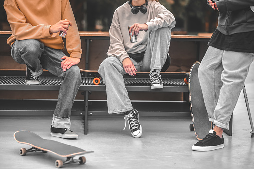Friends, communication. Three skateboarders in comfortable clothes with skateboards spending their free time chatting near bench, faces are not visible