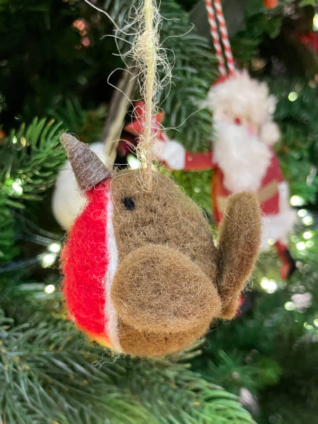 istock Close-up image of needle felt robin redbreast ornament hanging on artificial Christmas tree branch, spruce needles, blurred white fairy lights background 1352141609