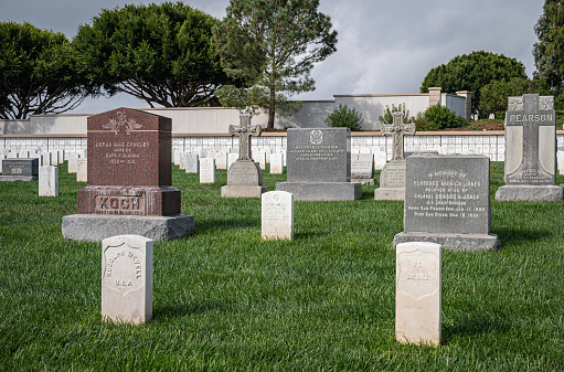 San Diego, California, USA - October 5, 2021: Fort Rosecrans National Cemetery. group of mixed style tombstones set among white simple graves with green foliage in back under gray cloudscape.