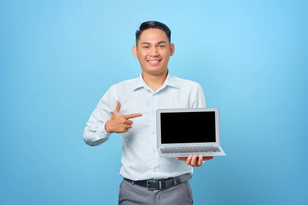 istock Smiling young handsome businessman pointing at a laptop with a blank screen on blue background 1352164420
