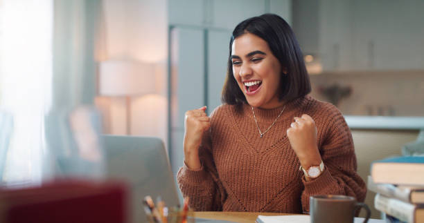 photo d’une jeune femme acclamant tout en utilisant un ordinateur portable pour étudier à la maison - joy cheerful happiness smiling photos et images de collection