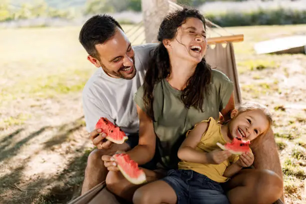 Photo of Cheerful family eating watermelon in hammock at the backyard.