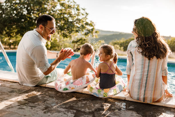 Happy family talking by the pool in summer day. Happy parents and their small kids talking while relaxing by the pool at the backyard. Copy space. back yard stock pictures, royalty-free photos & images