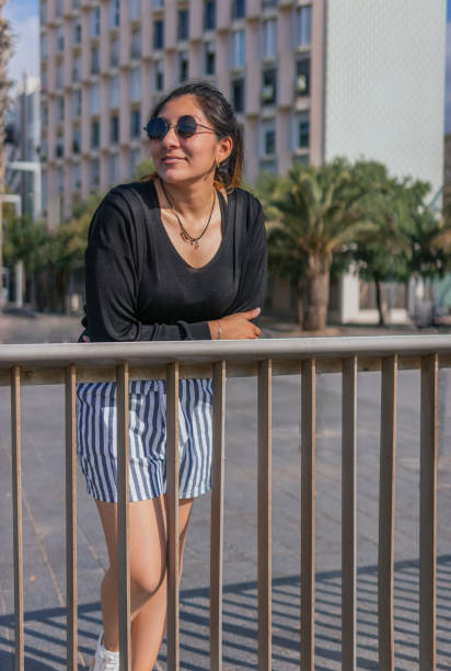 A latin woman on the street looking to the right Photo of a latin woman on the street resting her arms on a railing and looking to the right standing jeans shorts women latin american and hispanic ethnicity stock pictures, royalty-free photos & images