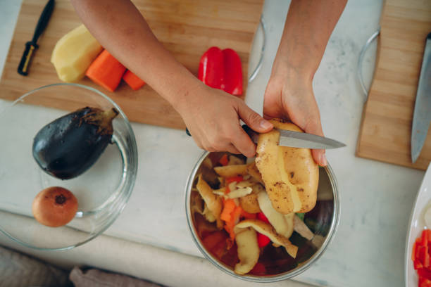 Woman's hand peeling a potato and putting a skin into a bowl for recycling.