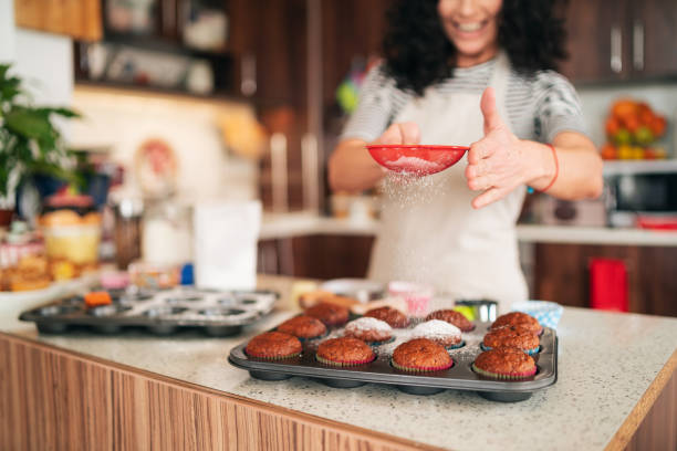 Curly brunette at home, using a strainer to sprinkle powdered sugar at freshly baked homemade cupcakes in her kitchen. Selective focus on her hands.