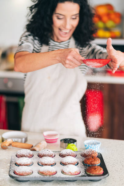 Curly brunette at home, using a strainer to sprinkle powdered sugar at freshly baked homemade cupcakes in her kitchen. Selective focus on her hands.
