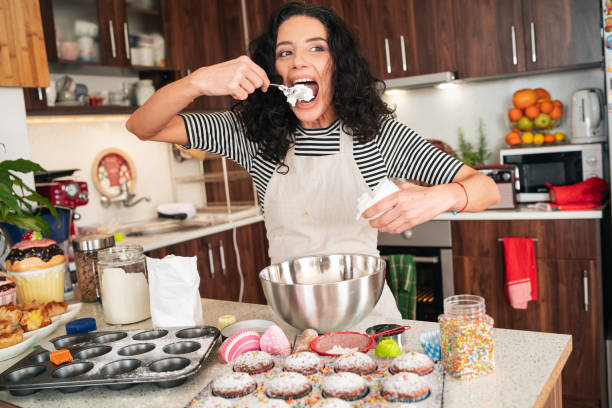 istock Beautiful brunette making cupcakes in her kitchen, making fun with sugar decoration and eating from the whipped cream with big spoon 1352221996