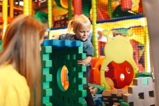 Happy Kid Playing With Big Puzzles in a Playground Center. Child With Young Mother Having Fun Together. Boy Contructing House With Toy Blocks