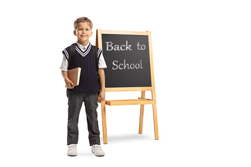 Schoolboy in a uniform holding a book and standing in front of a chalk board with text back to school isolated on white background