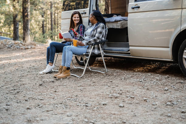 istock Multiracial women friends having fun camping with camper van while reading and drink coffee outdoor - Focus on african female face 1352229062