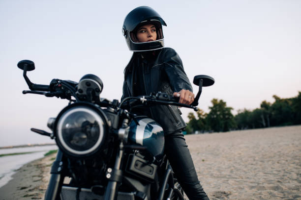 jovem senta-se em moto de capacete em praia perto do rio. - leather sensuality women jacket - fotografias e filmes do acervo