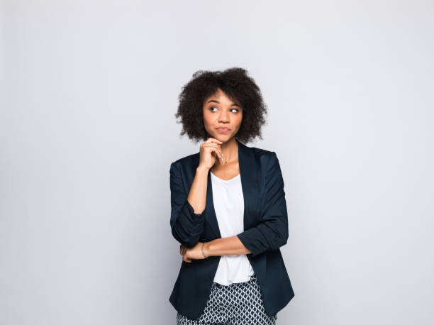 Portrait of thoughtful young businesswoman Portrait of beautiful young woman wearing navy blue jacket, standing with hand on chin and looking away. Studio shot of female entrepreneur against grey background. hand on chin stock pictures, royalty-free photos & images