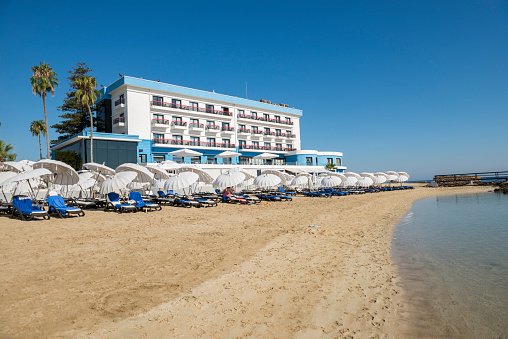 Famagusta, Cyprus - September 19, 2019: Arkin Palm Beach Hotel with beach umbrellas and tourists having sun bath near abandoned ghost town, Maras Famagusta, Northern Cyprus