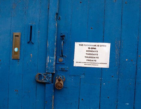 A sign attached to a blue wooden door indicating that the premises are used as a food bank which will be open on Mondays, Tuesdays, Thursdays and Fridays between the house of midday and two in the afternoon. Various locks, letterboxes and other door furniture are visible.