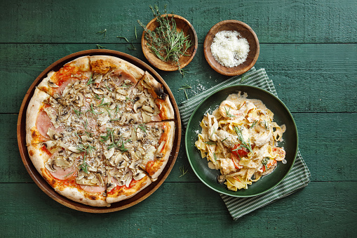 Man eating Italian fettuccine with prawns, salmon and herbs. Mushroom-prosciutto pizza. Flat lay top-down composition on dark green background.