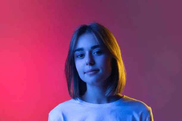 Stand at gaze. Close-up of young cute girl's face looking at camera isolated on pink-purple studio backgroud in neon. Human emotions, facial expression, youth fashion concept. Copy space for ad