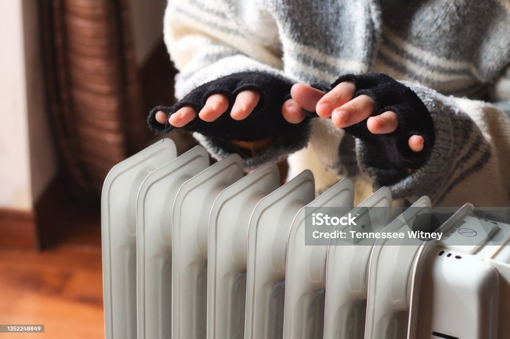 Person heating their hands at home over a domestic portable radiator in winter Radiator - Heater Stock Photo