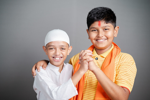 Happy Hindu Muslim kids showing unity by holding hands together by looking at camera on gray background - concept of diversity, bonding and multiethnic friendship