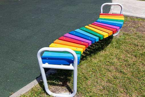 The curved wooden bench is painted in rainbow colors. An empty bench in a children's play area during the COVID pandemic