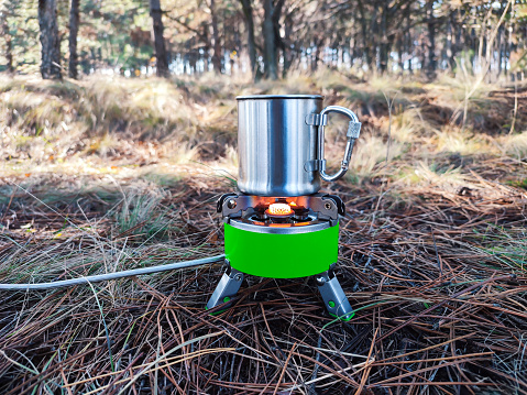 Metal cup of hot tea or coffee on portable gas burner or camping stove fire outdoor. Eco tourism and hiking concept