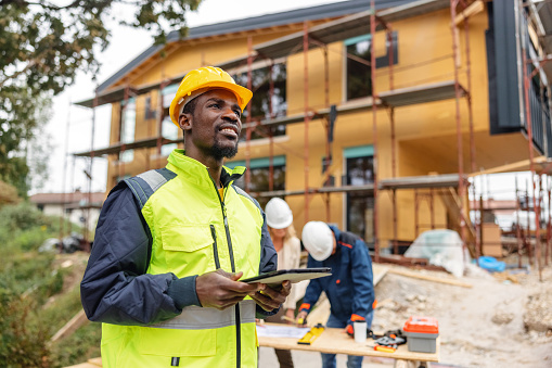 Front view of black male construction site manager wearing hard hat and high visibility vest, using his digital tablet while visually inspects a building project at construction site.
