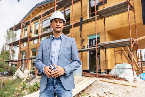 Portrait of confident chief engineering officer in mid 40s, wearing white hard hat, standing straight and holding smartphone at construction site.