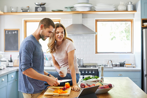 Smiling man chopping vegetables while woman reading recipe on digital tablet. Couple preparing food together at kitchen island. They are cooking at home during lockdown.