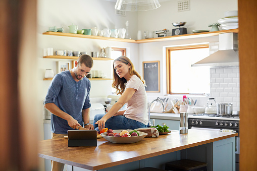 Woman pointing at digital tablet while sitting on kitchen island. Man is chopping vegetables while reading recipe. Couple is preparing food together in kitchen.