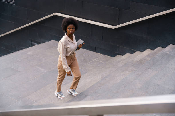 A dark-skinned girl in a beige classical outfit with afro hairs, ascending the concrete stairs A dark-skinned girl in a beige classical outfit with afro hairs, ascending the concrete stairs somewhere in a big city center, we see her from the high angle view in a daylight african american architecture stock pictures, royalty-free photos & images