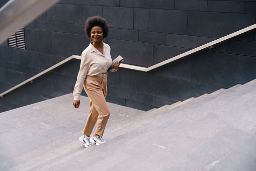 A smiling dark-skinned girl in a beige classical outfit with afro hairs, ascending the concrete stairs somewhere in a big city center, we see her from the high angle view in a daylight